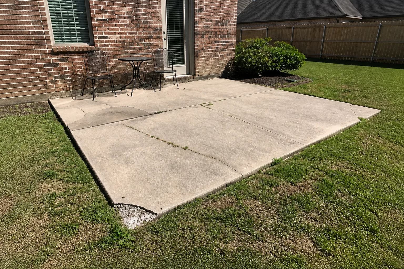 A cracked concrete patio outside a brick back door, with a metal table and chairs crowded against the wall in direct sun