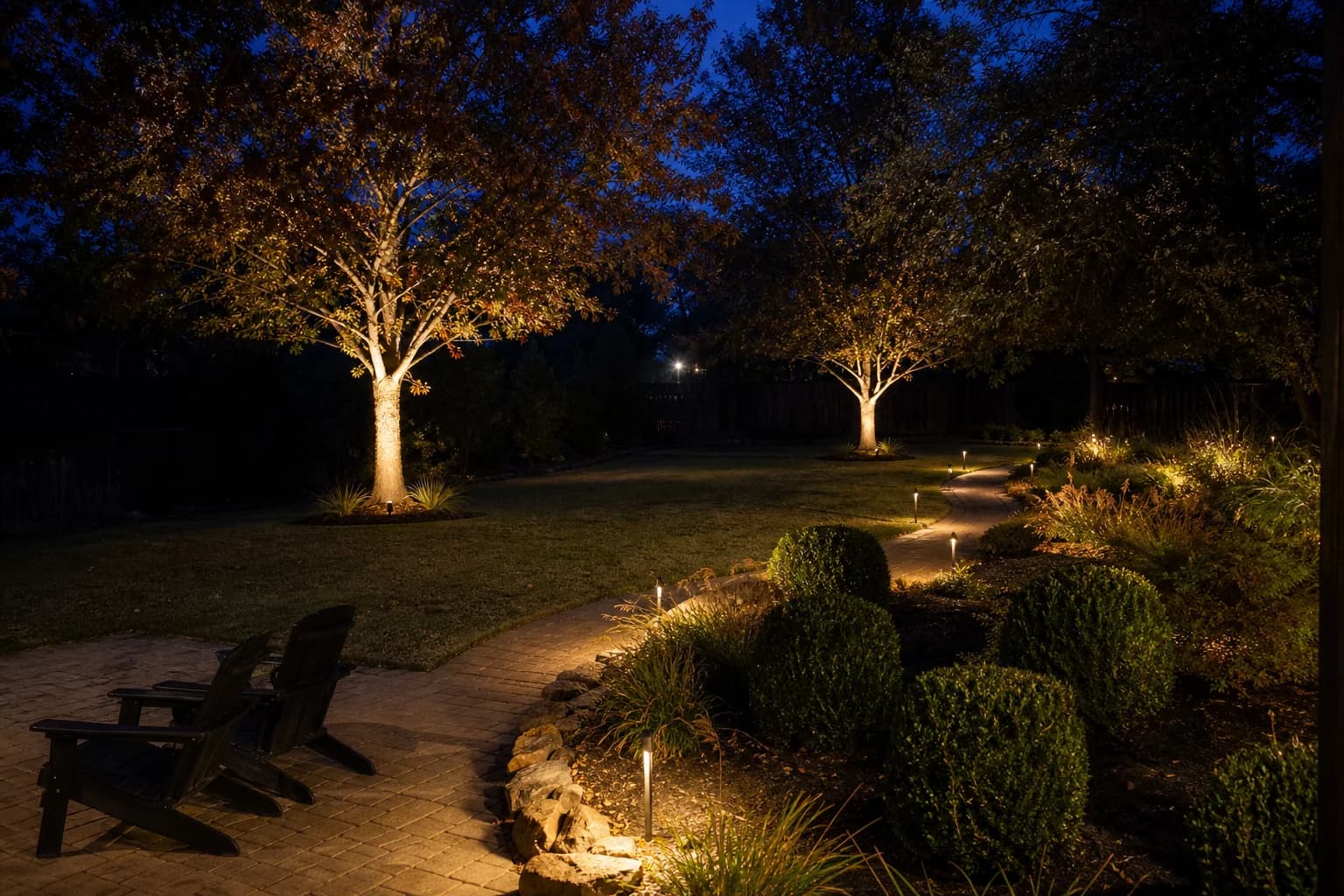 A backyard lit after dark, with path lights washing a curved walkway and two trees uplit from fixtures at their bases