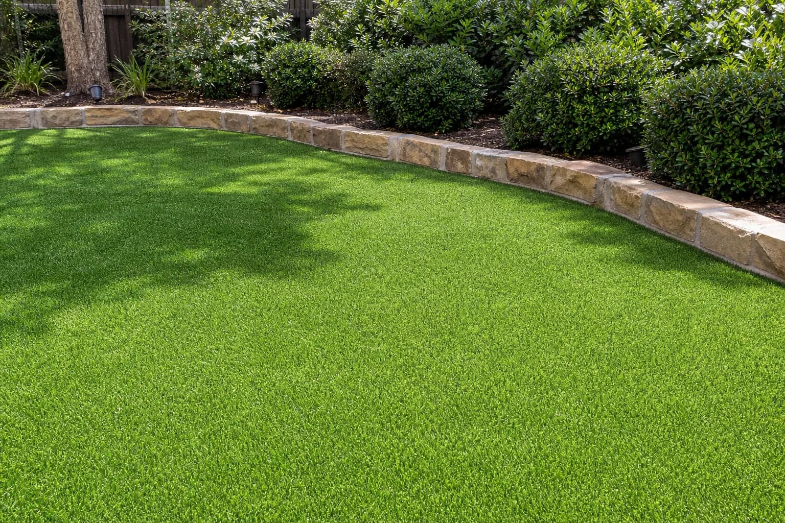 A synthetic turf lawn running edge to edge under dappled shade, meeting a curved limestone border with trimmed shrubs behind it