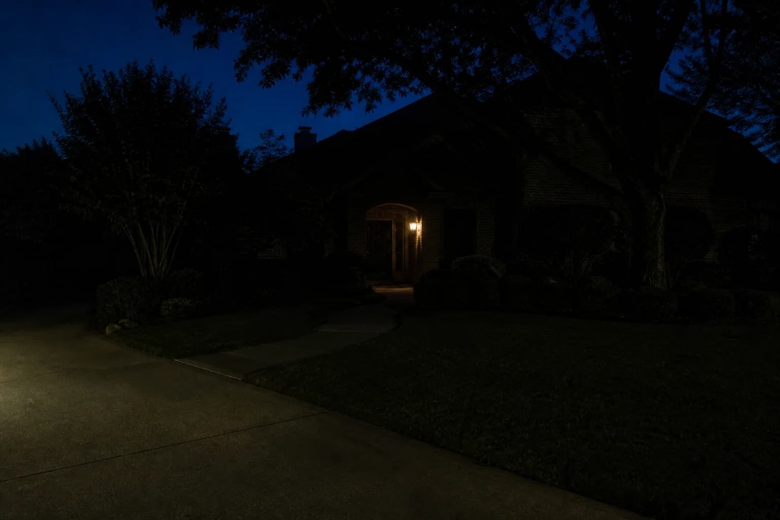 A front walkway and planting beds after dark, lost in shadow except for one porch light beside the door
