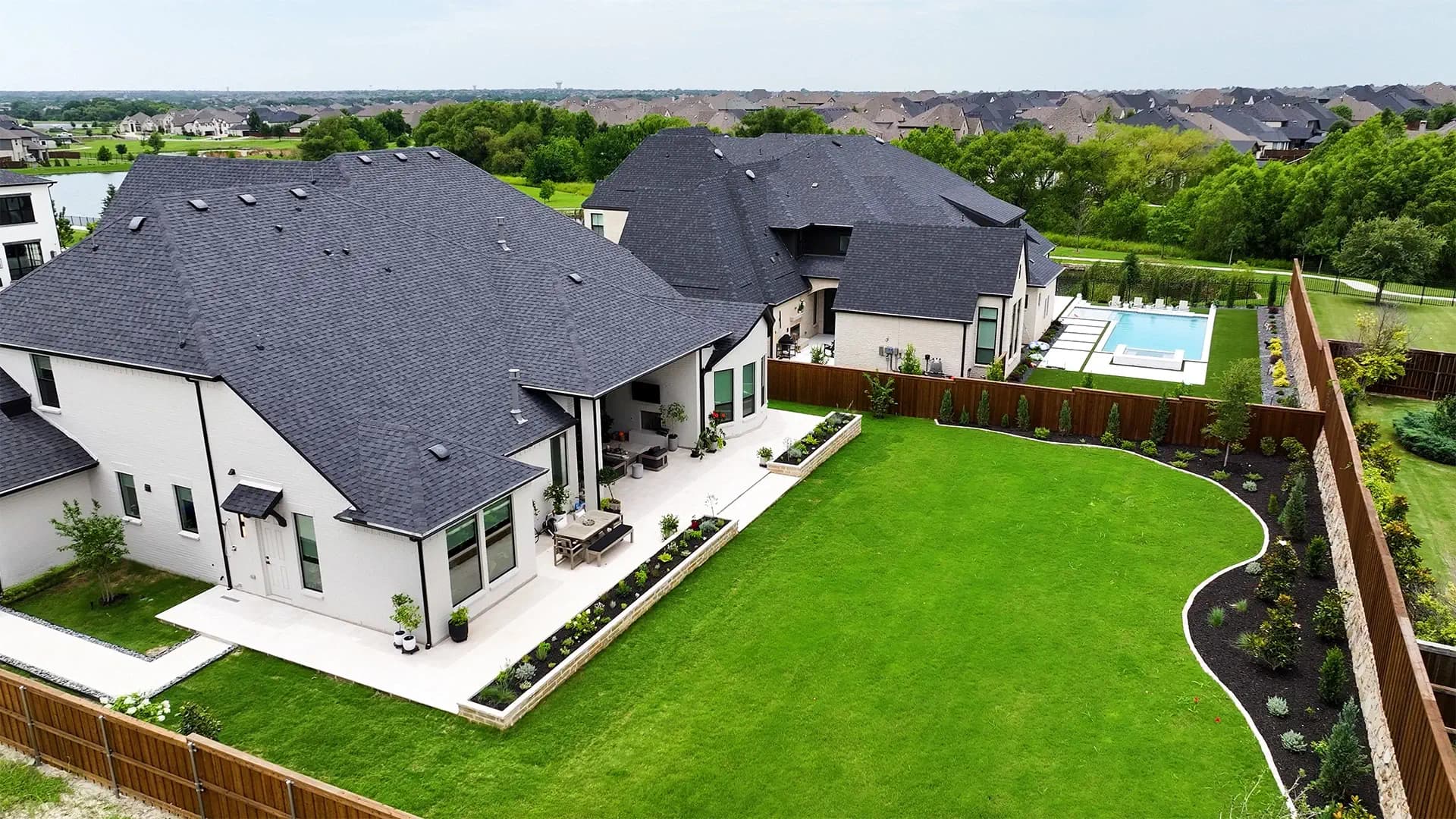 Aerial view of a completed backyard with a travertine patio, stone-edged planting beds, and a line of young screening trees along the fence