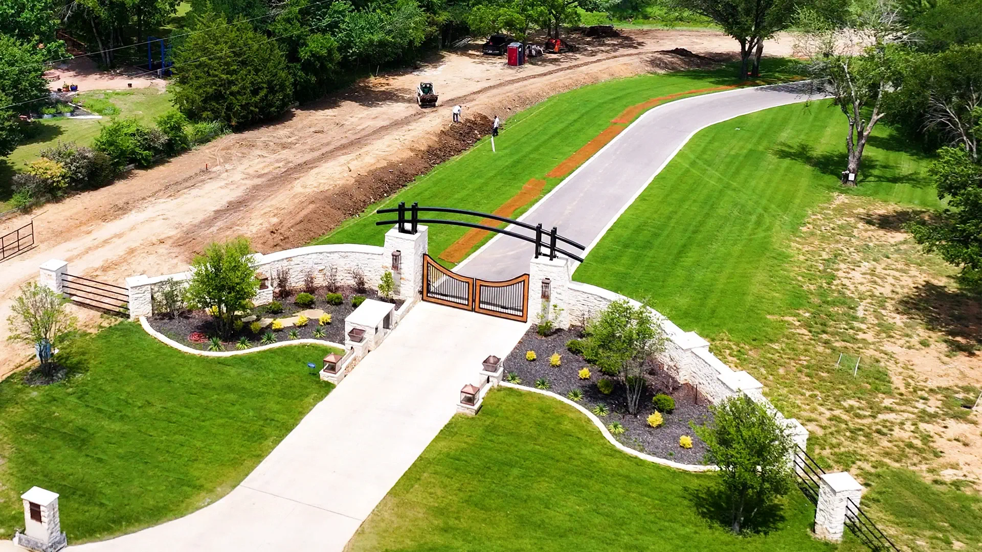 A custom estate entrance in Colleyville with curved limestone walls, an arched iron gate, and new landscaping along the driveway