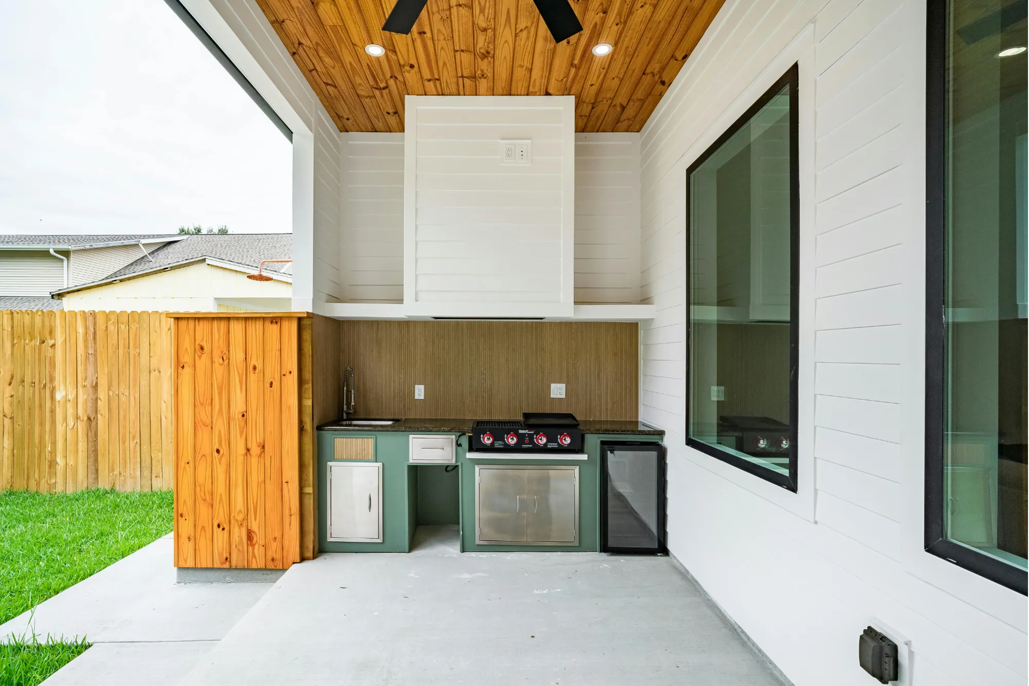 A built-in outdoor kitchen under a covered patio with a grill, sink, beverage fridge, and stone countertop